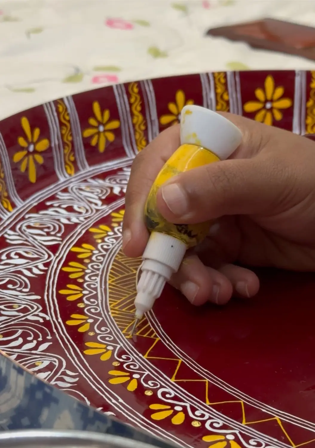 Hands painting yellow floral Aipan motifs onto a deep red plate with a fine-tipped cone