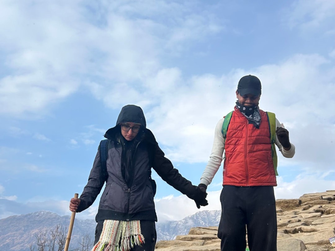 Two trekkers holding hands on the rocky Tungnath–Chandrashila trail with Himalayan peaks behind