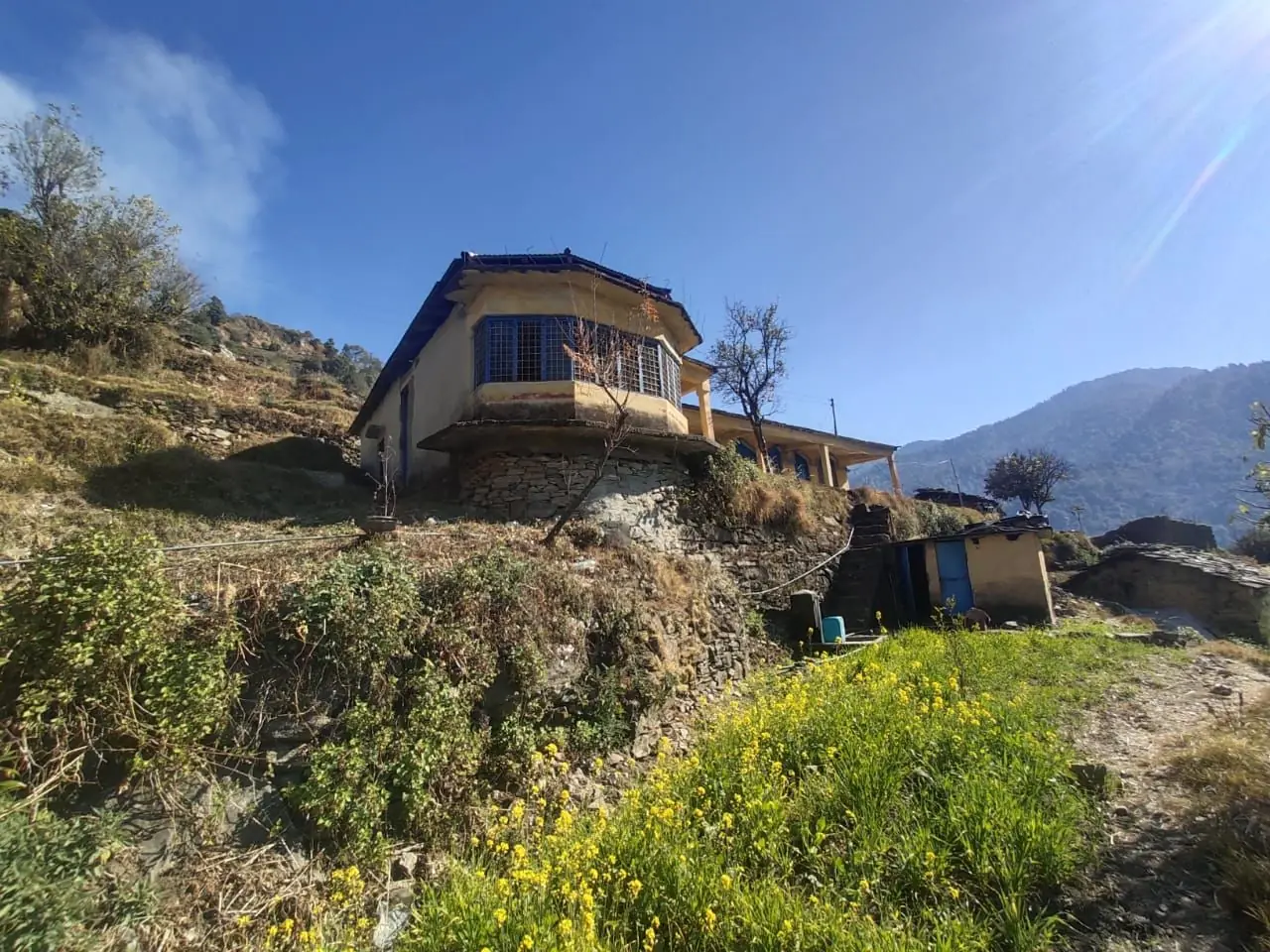 The original yellow mountain building from below, with mustard flowers in the foreground and the Himalayan slope behind it