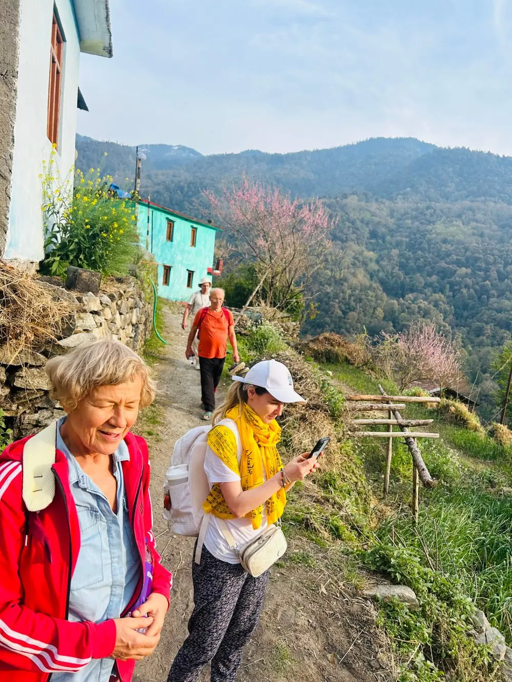 Guests pausing on the village path, with painted village houses, blossoming trees and forested ridges beyond