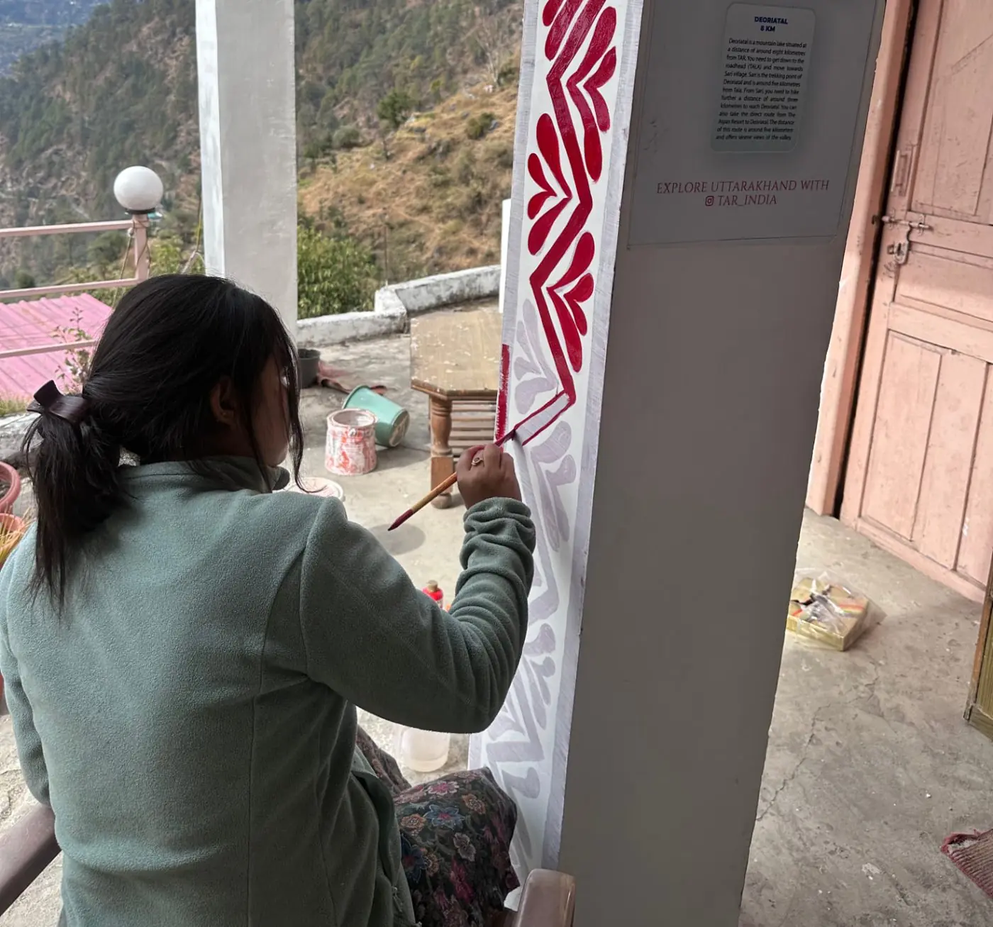 An artist painting a red Aipan border onto a pillar on the terrace, with the Chopta valley behind