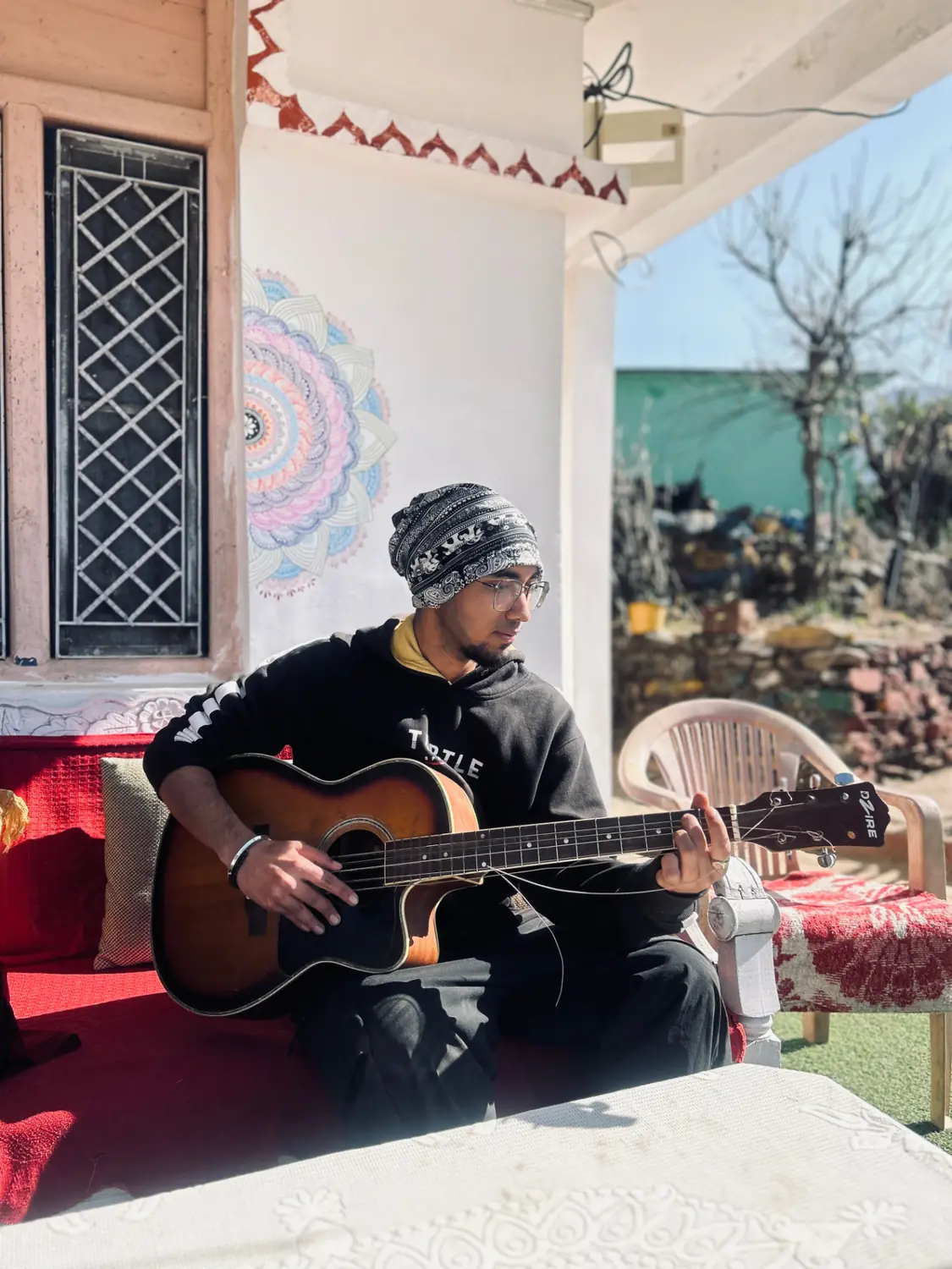 A guest playing acoustic guitar in the sun-drenched courtyard with Aipan art on the wall